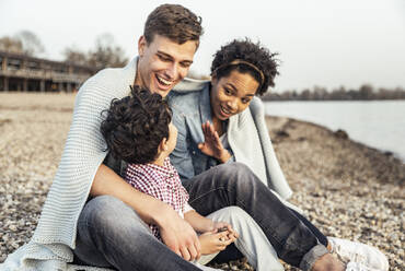 Parents smiling at son while sitting covered in blanket over pebble - UUF23013