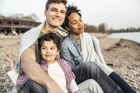 Smiling man sitting with family covered in blanket over pebble - UUF23014