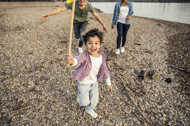 Playful boy holding sticks while playing with parents in background over pebble - UUF23016