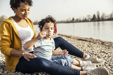 Boy blowing in bubble wand while sitting with woman on pebble - UUF23026