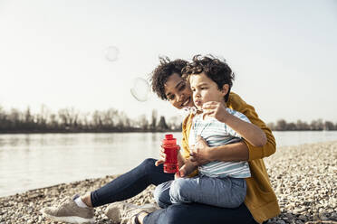 Junge spielt mit einem Seifenblasenstab auf dem Schoß seiner Mutter an einem sonnigen Tag - UUF23027