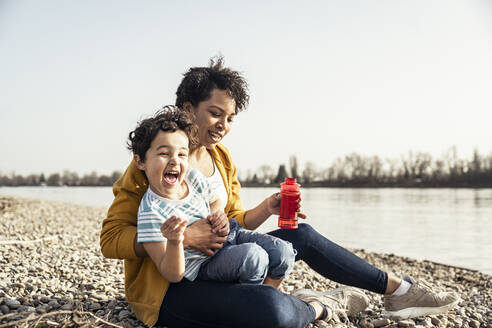 Cheerful boy with bubble wand sitting on mother's lap over pebble - UUF23028