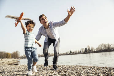 Playful man and boy holding airplane toy while running by lakeshore on sunny day - UUF23041