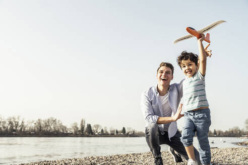 Playful boy playing with airplane toy by father crouching on pebbles - UUF23045