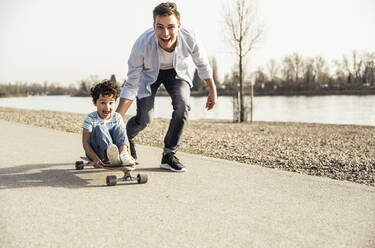 Cheerful man and boy playing with skateboard on road during sunny day - UUF23048