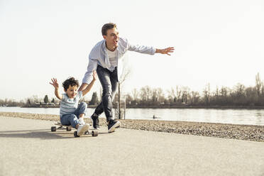 Vater läuft und schiebt seinen Sohn, der auf einem Skateboard sitzt, an einem sonnigen Tag - UUF23049