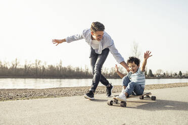 Junger Mann spielt auf einem Skateboard sitzend an einem sonnigen Tag - UUF23050