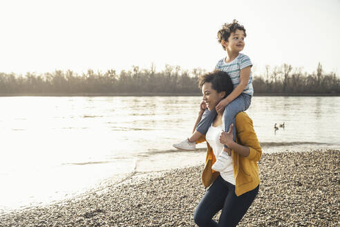 Junge Frau trägt Jungen auf den Schultern beim Laufen am Seeufer - UUF23054