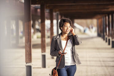 Smiling young woman with mobile listening music through in-ear headphones on sunny day - UUF23066