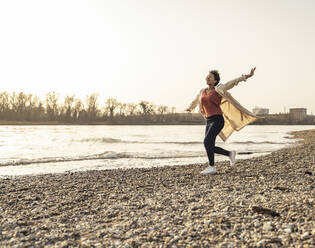 Cheerful woman wearing overcoat running by lake on sunset - UUF23074