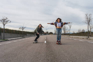 Female friends skateboarding on road against sky - RSGF00586