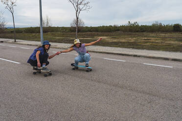 Playful female friends holding hands while crouching on skateboarding - RSGF00603