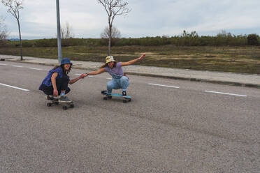 Verspielte Freundinnen halten sich an den Händen, während sie auf dem Skateboard hocken - RSGF00603