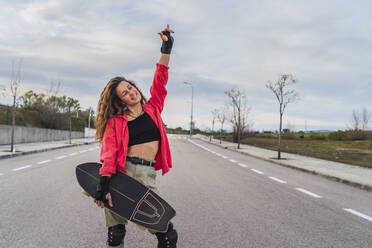 Lächelnde Frau mit Skateboard gestikuliert Horn Zeichen, während auf der Straße gegen den Himmel stehen - RSGF00608