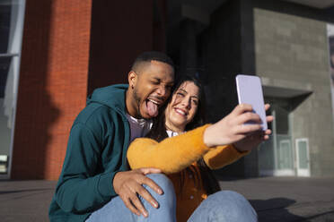 Junger Mann macht ein Gesicht, während seine Freundin ein Selfie mit dem Smartphone macht - MTBF00909