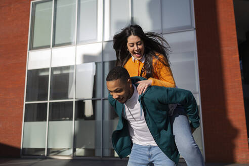 Mann nimmt seine fröhliche Freundin vor einem modernen Gebäude huckepack - MTBF00911