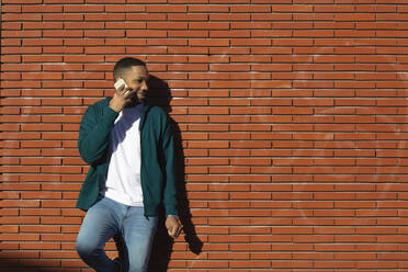 Lächelnder junger Mann, der sich an die Wand lehnt und mit seinem Handy spricht - MTBF00915