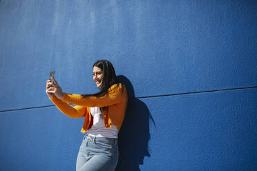 Smiling woman taking selfie through mobile phone against blue wall - MTBF00927