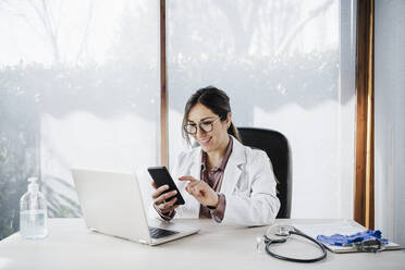 Lächelnde Ärztin, die ein Mobiltelefon benutzt, während sie am Schreibtisch im Krankenhaus sitzt - EBBF02649