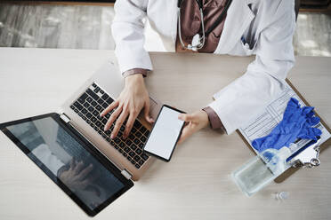 Eine Ärztin hält ein Smartphone in der Hand, während sie einen Laptop am Schreibtisch benutzt - EBBF02651