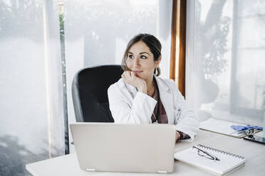 Smiling female medical professional with hand on chin day dreaming while sitting at desk - EBBF02658