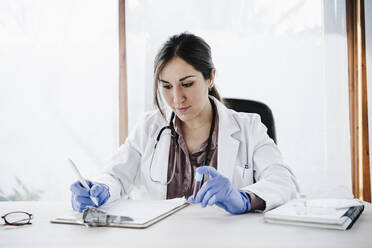Female medical expertise writing blood test reports while holding blood sample at desk - EBBF02672