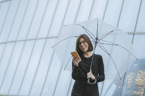 Cheerful young woman with umbrella using smart phone while standing in front of glass building - MTBF00930