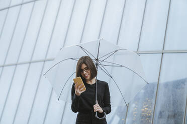 Lächelnde Frau, die ein Mobiltelefon benutzt und einen Regenschirm gegen ein Glasgebäude hält - MTBF00931