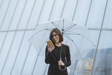 Fröhliche junge Frau, die einen Regenschirm hält und ein Smartphone vor einem Glasgebäude benutzt - MTBF00932