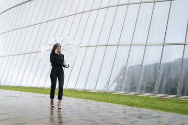 Junge Frau mit Regenschirm, die mit ihrem Handy telefoniert, während sie auf dem Fußweg neben einem Glasgebäude spazieren geht - MTBF00933