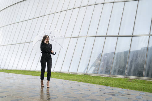 Young woman with umbrella talking on mobile phone while walking on footpath by glass building - MTBF00933