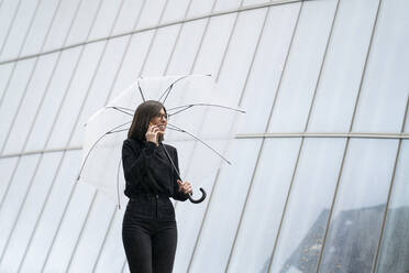 Lächelnde Frau, die einen Regenschirm hält, während sie bei einem Glasgebäude telefoniert - MTBF00934