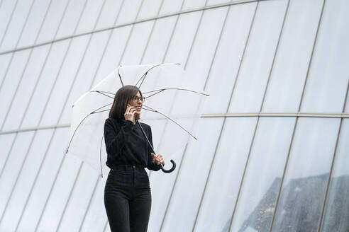 Smiling woman holding umbrella while talking on phone by glass building - MTBF00934