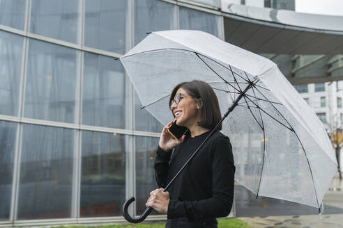 Fröhliche junge Frau, die einen Regenschirm hält, während sie bei Regen an einem Glasgebäude telefoniert - MTBF00936