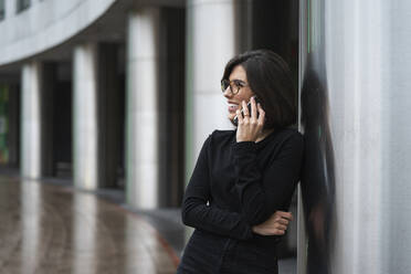 Lächelnde junge Frau, die sich an eine Säule lehnt, während sie mit einem Mobiltelefon spricht - MTBF00940
