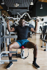 Männlicher Sportler sitzt auf einem Trainingsgerät im Fitnessstudio - EBBF02692