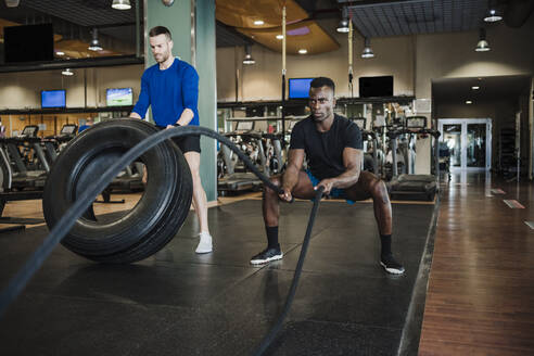 Männliche Sportlerfreunde trainieren mit Seil und Reifen im Fitnessstudio - EBBF02731