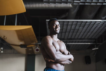 Shirtless sportsman smiling with arms crossed in gym - EBBF02791