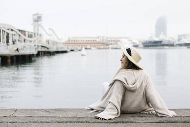 Junge Frau mit Hut, die sich auf einer Stützmauer am Wasser ausruht - XLGF01313