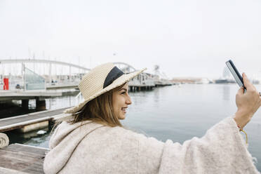 Smiling young woman with hat taking selfie through mobile phone at the waterfront - XLGF01315