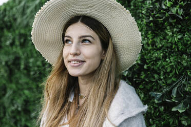 Thoughtful young woman in hat by green plant wall - XLGF01319