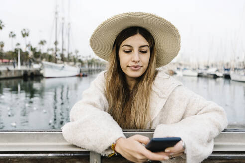 Junge Frau mit Hut benutzt ein Mobiltelefon, während sie am Hafen steht - XLGF01321