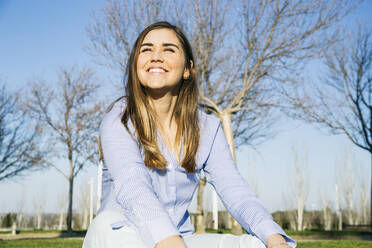 Glückliche Frau lächelt, während sie an einem sonnigen Tag im Park sitzt - ABZF03489