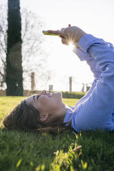 Glückliche Frau, die ein Mobiltelefon benutzt, während sie an einem sonnigen Tag im Gras liegt - ABZF03491