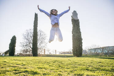 Carefree woman jumping with arms outstretched at park - ABZF03494