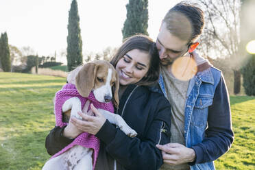 Pet owners looking at dog in sweater while standing at park - ABZF03499