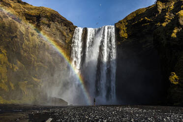 Unerkennbarer Reisender in der Nähe eines beeindruckenden Wasserfalls und eines Regenbogens in den Bergen während eines Urlaubs in Island - ADSF21433
