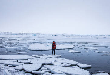 Rückenansicht eines anonymen Forschers in warmer Kleidung, der auf einem Stück Eis im Meer nahe der Küste steht und die winterliche Meereslandschaft in Island bewundert - ADSF21438