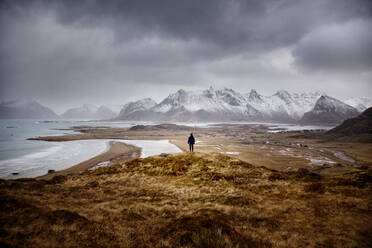 Rückenansicht eines anonymen Forschers, der auf einem trockenen Hügel steht und die Aussicht auf schneebedeckte Berge und das gefrorene Wasser des Meeres an einem bewölkten Wintertag in Norwegen bewundert - ADSF21444