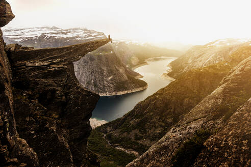 Fernblick auf einen Forscher, der bei Sonnenuntergang am Rande der Trolltunga-Felsformation sitzt, während eines Urlaubs in Norwegen - ADSF21448